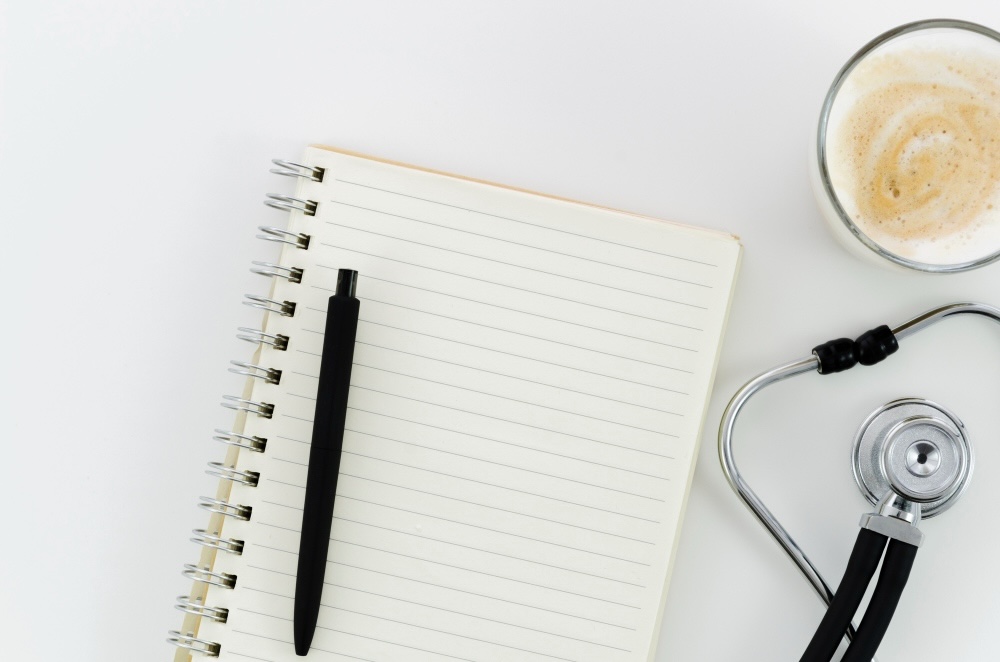 A pen, notepad, stethoscope, and coffee on a white counter.